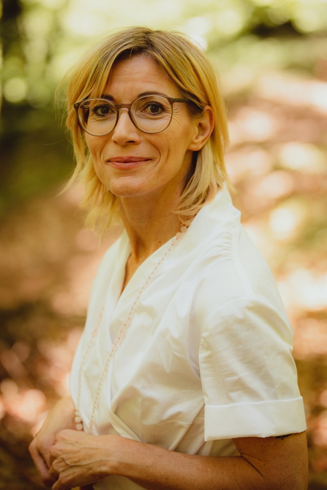 Das Bild zeigt eine Frau mit Brille und schulterlangen blonden Haaren und weißem Oberteil mit Ärmeln in der Natur. Sie lächelt in die Kamera. Die Stimmung im Hintergrund lässt sich erahnen, dass sie an einem sonnigen Tag im Wald steht.