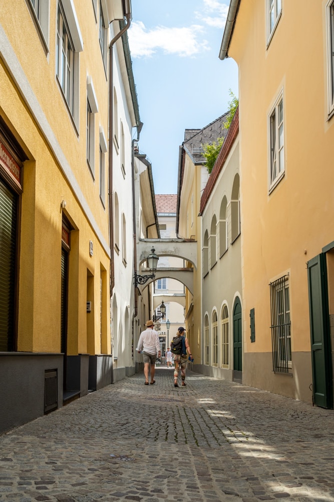 Die Badgasse nach Süden Fotografiert - die Häuserfassade links ist von der Sonne beschienen, die rechte liegt im Schatten, am Boden treffen keine Sonnenstrahlen auf in der engen Gasse, ein Pärchen flaniert durch die Gasse, der Himmel ist blau mit ein paar Schleierwolken, es ist ein warmer Sommertag