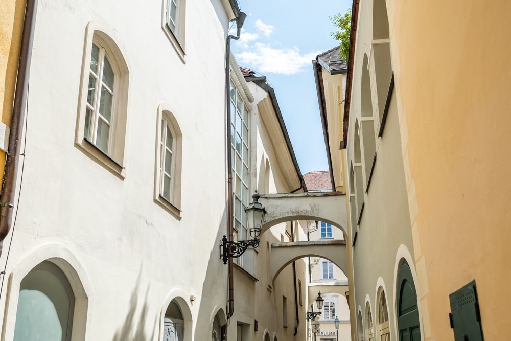 Die Badgasse Klagenfurt im Spätsommer - man sieht von unten nach schräg oben fotografiert die Häuserfassaden der Gasse und die Rundbögen, die beide Seiten verbinden, die linke Häuserfassade ist von der Sonne beschienen, die rechte liegt im Schatten, der Himmel ist blau