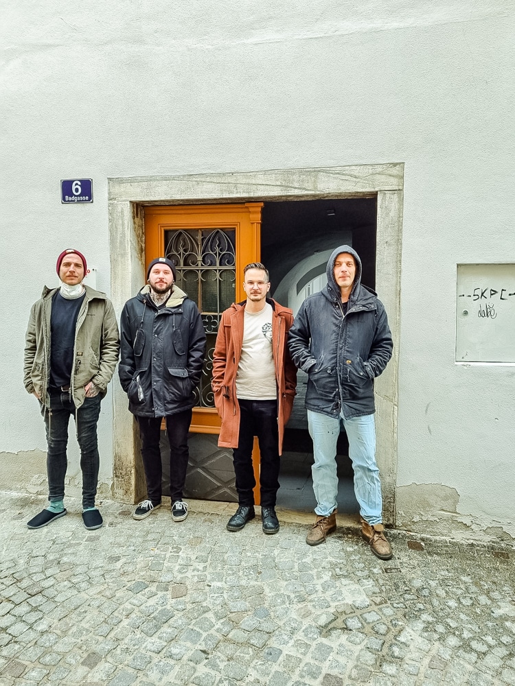 COTU - Lukas Schneider, Daniel Bosnjak, René Znidar und Gabriel Schneider stehen nebeneinander vor einer doppelflügeligen Holztür mit Glaseinsatz, die vier Männer tragen Winterjacken und drei und Hauben, der Boden ist gepflastert, an der weißen Wand ist links ober der Tür, die mit Steinplatten gerahmt ist, das Adressschild "Badgasse 6", rechts neben der Tür ist ein kleiner Kasten in die Mauer eingelassen, die ebenso weiße Tür ist mit Graffiti beschmiert. Die Farbe der Mauer ist in Bodennähe abgeblättert., der rechte Türflügel ist offen, man sieht in den dunklen Gang, der in einem Innenhof führt