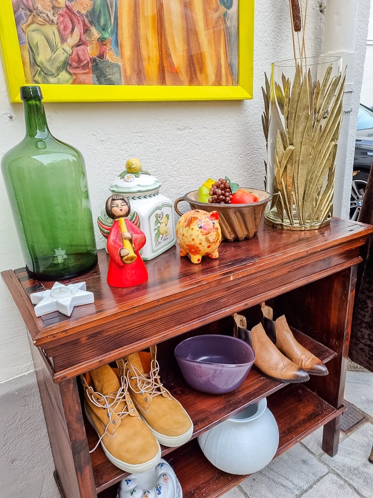 Das Geschäft "Fundus" in der Badgasse - am Bild ein kleines Holzregal mit unterschiedlichen Produkten: Tonfiguren, Vasen, Schüsseln, Schuhen, Teelichter. Über dem Regal, das an einer weißen Wand steht, erkennt man das untere Drittel eines gemaltes Bild in einem gelben Rahmen