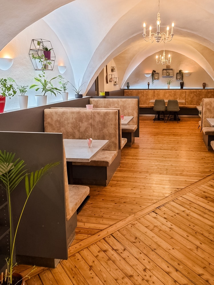 Im SU vegetarisches Restaurant ist ein Holzboden und ein weißes Gewölbe, von der Decke hängen kleine weiße Kronleuchter, auf der Fensterbank links stehen ein paar Pflanzen, Links der Wand entlang, sowie auf der Rückseite und leicht zu erkennen auch rechts am Bild sind mehrere Tische für zwei bis vier Personen, die stoffbezogenen Bänke haben hohe Lehnen, in der Mitte ist ein breiter Gang.