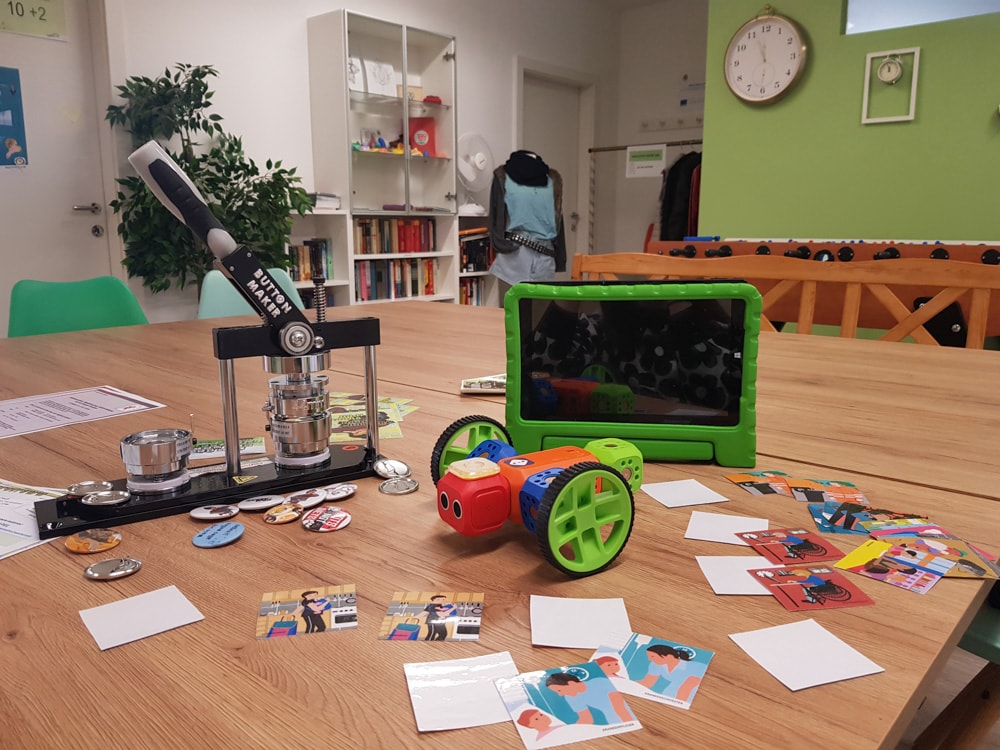 Forschen und experimentieren mit EqualiZ-Gemeinsam vielfältig - in einem Raum mit grünen und weißen Wänden sind auf einem Tisch ein kleiner Roboter mit Rädern, ein Tablet, ein Button-Maker und viele Arbeitskärtchen gelegt
