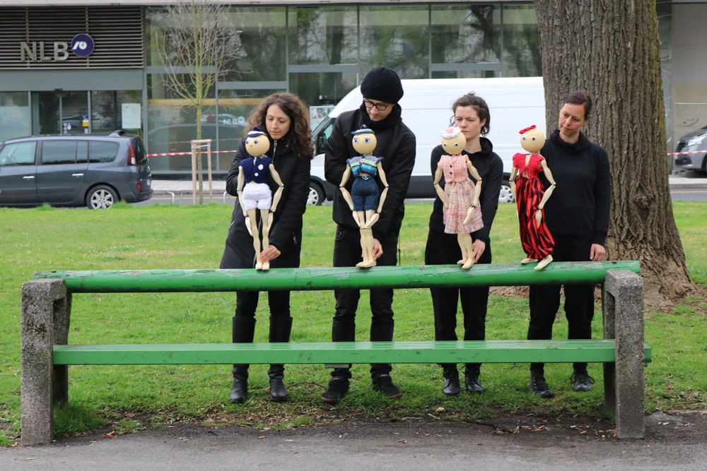 Theater Strick - drei Frauen und ein Mann in schwarz gekleidet halten jeweils ein e Handpuppe, die Personen stehen hinter einer Holzparkbank in grün mit Betonsäulen, die Puppen stehen auf dem Balken - die Lehne von der Bank. Im Hintergrund steht ein großer Baum, dahinter ist ein Gebäude und Autos