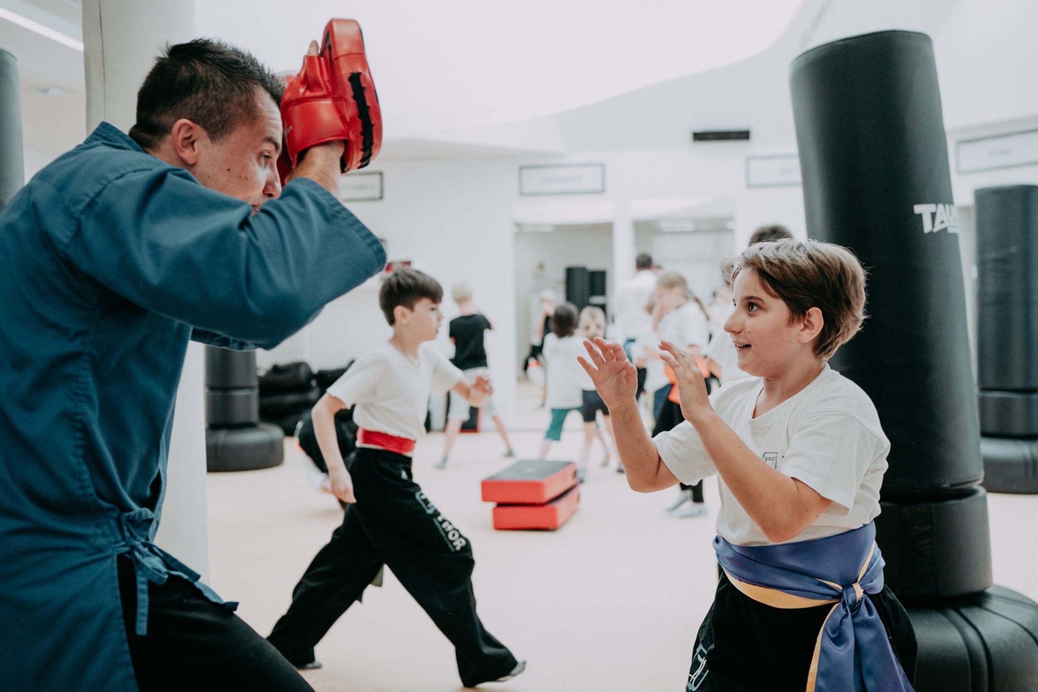 THOR Training - beim Kampfkunsttraining lernen Kinder auch ihr Selbstbewusstsein zu stärken - am Bild ist der Trainer links mit türkisem Oberteil und schwarzer Hose in Deckungshaltung, er hält seine Hände mit roten gepolsterten Handschuhen vor sein Gesicht. Vor ihm ist ein Bub der sich bereit macht auf die roten Handschuhe zu schlagen. Er trägt ein weißes T-Shirt, eine schwarze Hose und ein blau-gelbes Band als Gürtel. Im Hintergrund sieht man verschwommen noch andere Kinder und Boxsäulen.