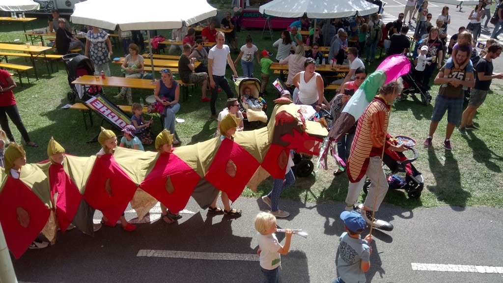 Auf einem Fest für Familien sind im Hintergrund auf einer Wiese Bierbänke aufgestellt, auf welchen Kinder und ihre Eltern sich tummeln, im Vordergrund spaziert auf einer Straße ein Clown mit riesigen Schuhen vor einem "Zug" von Kindern unter einem Drachenkostüm. Ein Kind ist unter dem Kopf des Drachen geschlüpft, weitere Kinder sind dahinter und halten mit ihren Schultern den Körper des Drachens, die Kinder tragen auf ihren Köpfen Kappen, die die Schuppen des Drachens symbolisieren - das Kostüm ist von Jakotopia