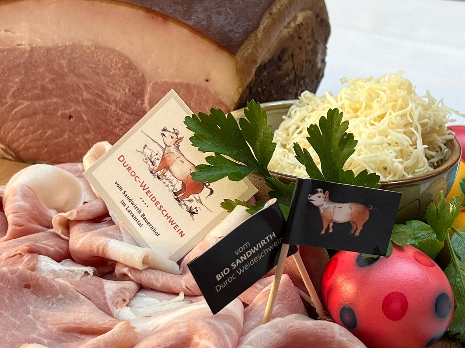 Eine Nahaufnahme von einer klassischen Osterjause in Kärnten mit aufgeschnittenem Osterschinken vorne links, rechts daneben ein rotes Osterei mit blauen und dunkelroten Punkten, hinter dem Ei in einer Schüssel aufgeriebener Kren, frische Petersilie als Deko beim Ei dabei, hinten links ist der Schinken als ganzes zu sehen. Mit zwei Fähnchen und einer quadratischen Karte erkennt man am Bild, dass der Schinken vom Sandwirth ist. Es ist ein illustriertes Schwein abgebildet. Auf der Karte steht "Duroc-Weideschwein vom Sandwirth Bauernhof im Lavanttal".