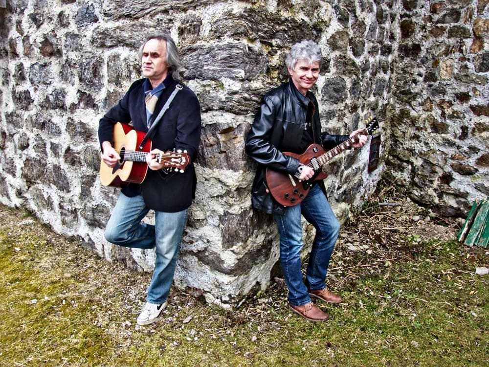 zwei Musiker mit Gitarre stehen an eine Steinwand gelehnt