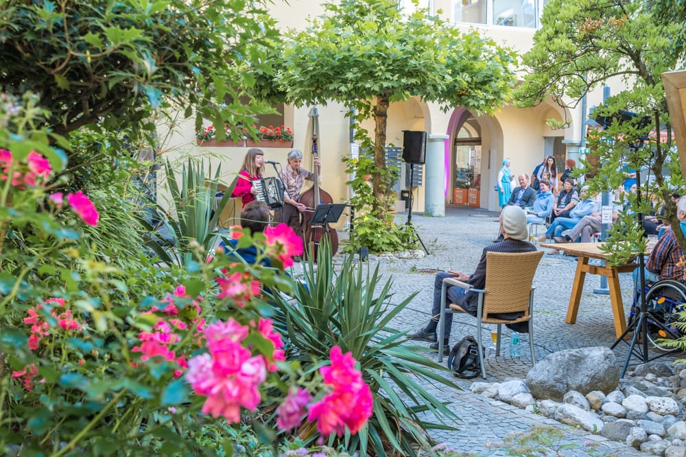 DonnerSzenen im Hof vom Stadtcafé Diakonie de la Tour, links unten im Vordergrund ist ein Strauch mit pinken Blüten, dahinter stehen zwei Künstler*innen, die zum Publikum gewandt Gitarre und Ziehharmonika spielen. Das Publikum sitzt auf Stühlen am rechten Bildrand, im Hintergrund sind Bäume und eine Häuserfassade mit Arkaden zu sehen