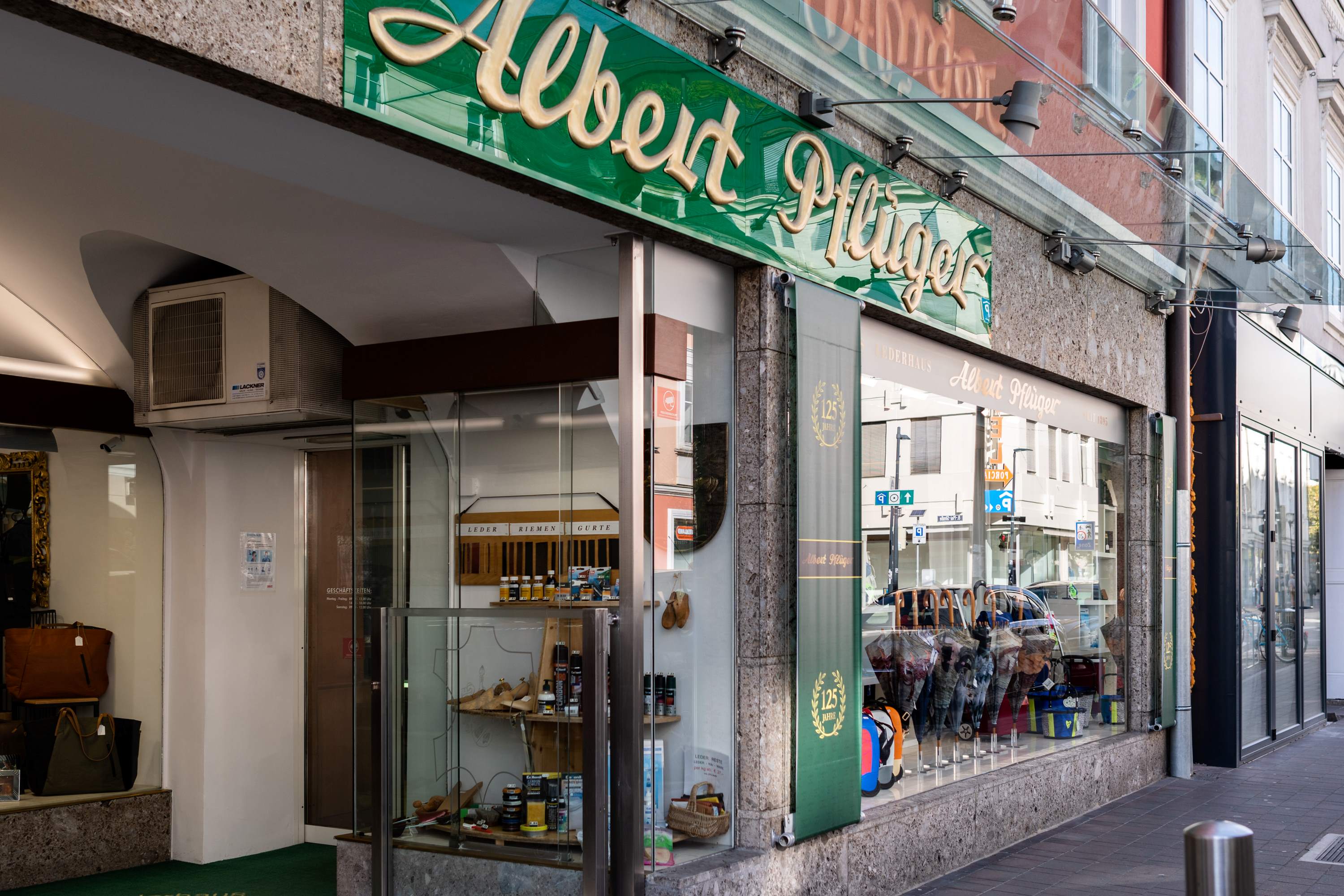 Der Eingangsbereich des Lederhaus Pflüger mit dem großen grünen Firmenlogo "Albert Pflüger" oberhalb der Tür, daneben Schaufenster