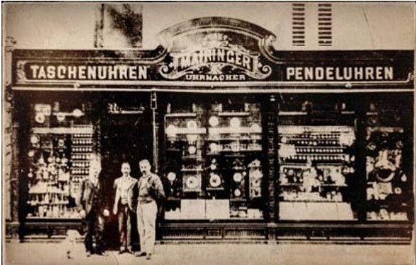 Eine Aufnahme des Geschäfts von Uhrmacher Mairinger aus der Zeit um die Jahrhundertwende, auf der drei Männer und ein Hund vorm Eingang stehen