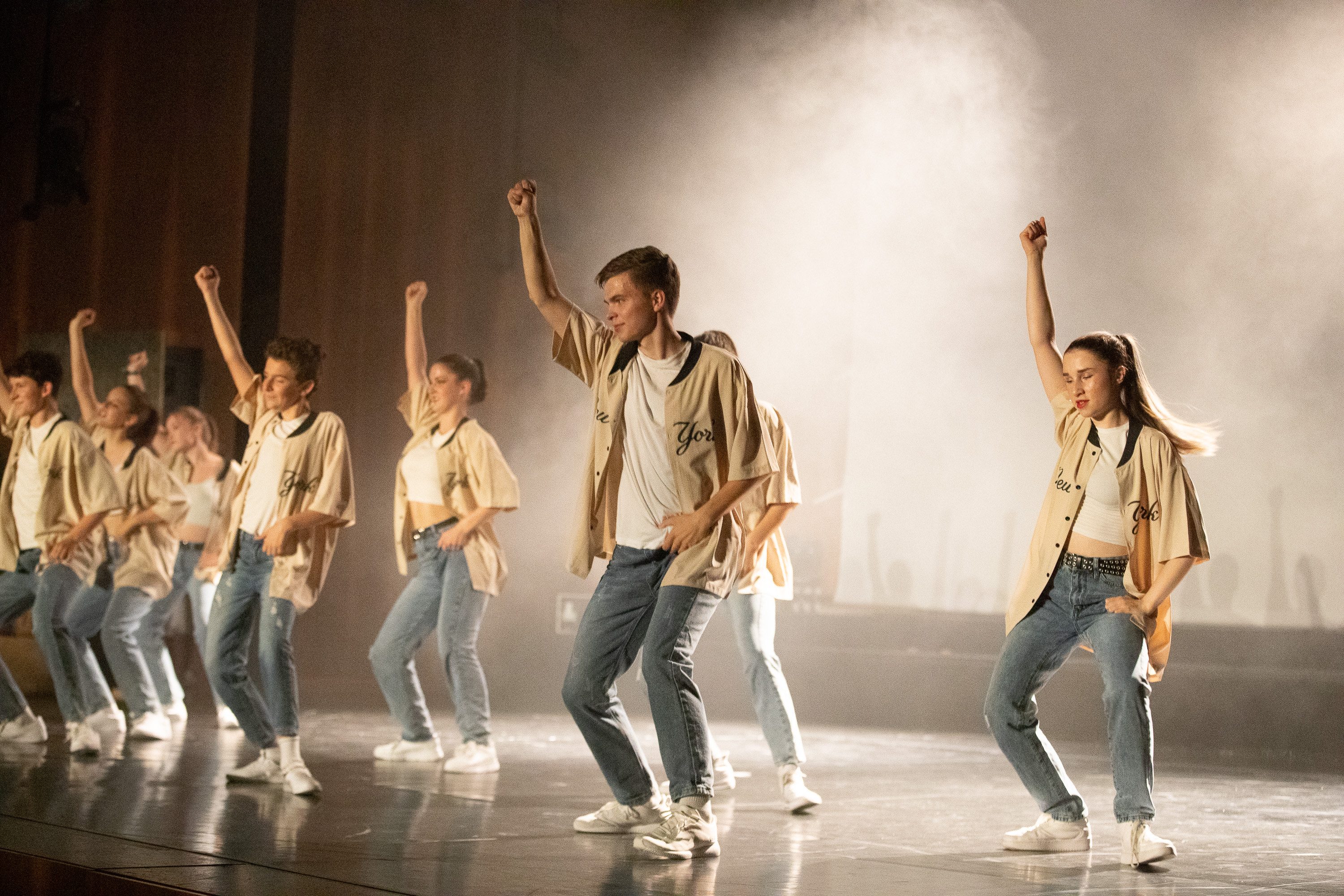 Tanzgruppe vom Tanzstudio SHINE Klagenfurt auf der Bühne, alle tragen blaue Jeans, ein weißes T-Shirt und ein offenes Baseball-Hemd in beige mit schwarzer Schrift "New York", weiße Schuhe und weiße Socken, die Bühne ist leicht vernebelt,