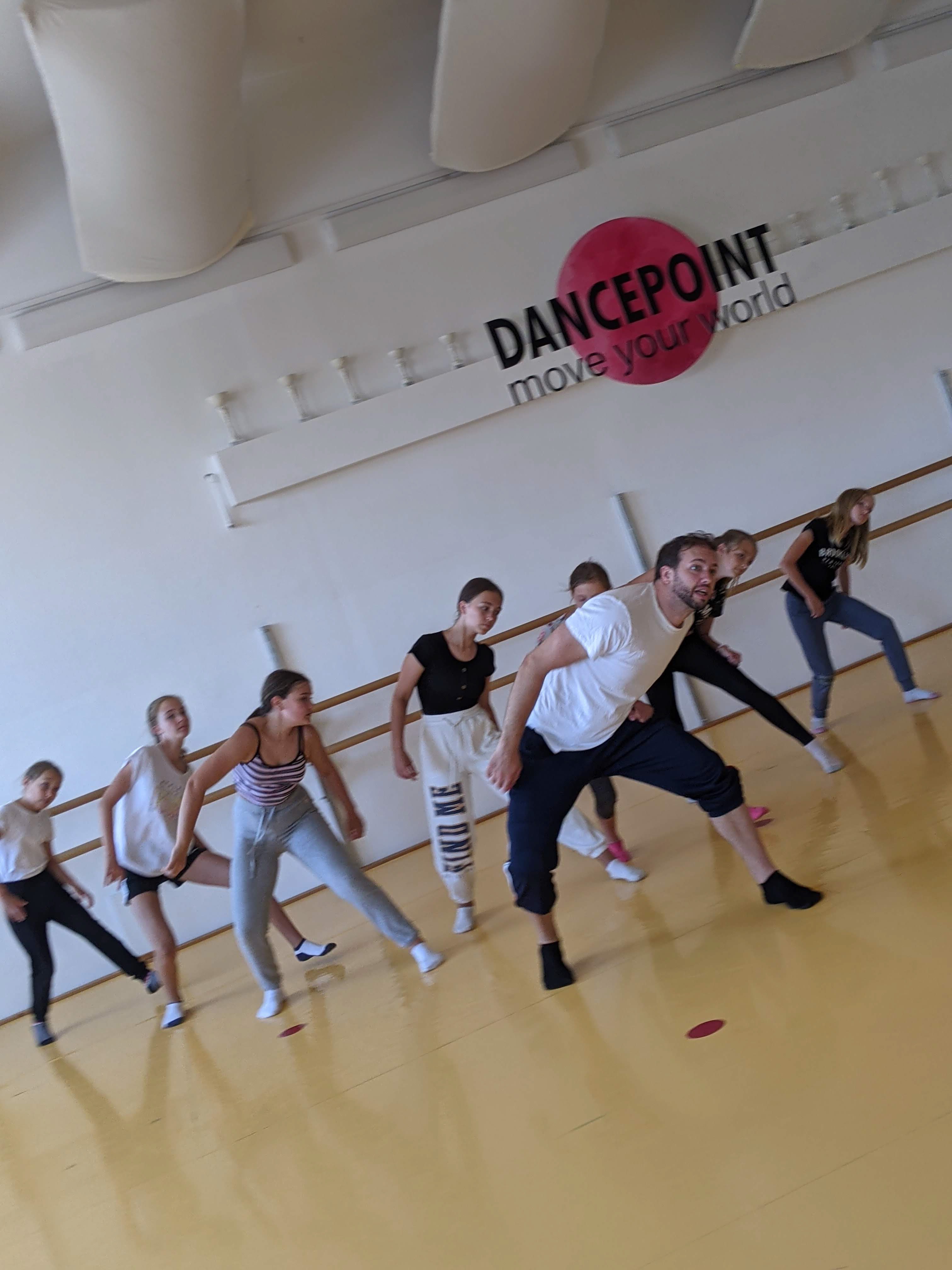 Eine Gruppe von Dancepoint mit Trainer im Studio beim Training, auf der Wand im Hintergrund ist das Logo angebracht "Dancepoint move your world" mit dem markanten roten Punkt in der Mitte