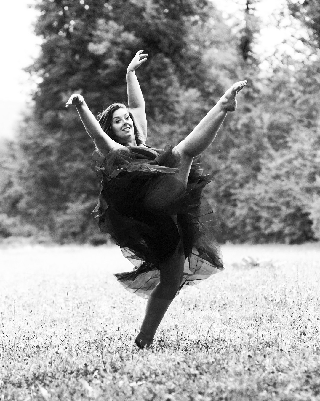 Tänzerin vom Tanzstudio SHINE in einem Park, sie trägt ein schwarzes Kleid, beide Arme sind nach oben gestreckt und ein Bein auf Kopfhöhe in der Luft, die Zehen gestreckt, das linke Bein am Boden ist leicht gebeugt, der Oberkörper ist leicht nach hinten gelehnt, der Blick geht nach vorne, schwarz-weiß-Aufnahme