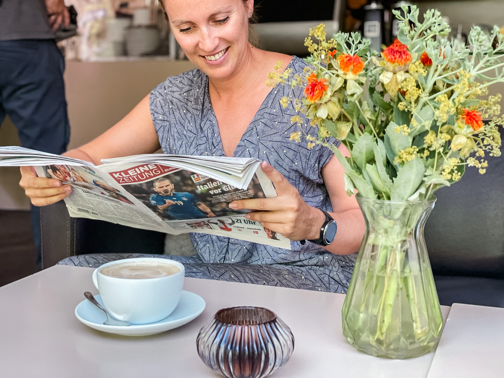 Junge Frau liest lächelnd in der Kleinen Zeitung und sitzt vor einem großen Cappuccino und einem Strauß Blumen im Como in der Bahnhoftstraße in Klagenfurt
