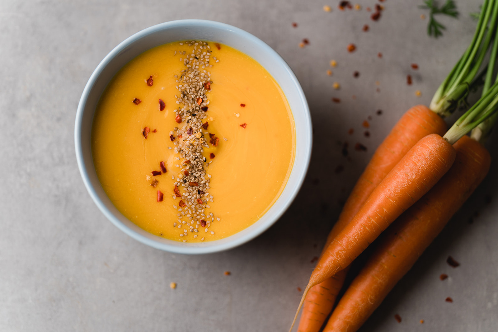 Ansicht von oben auf ein Teller mit Karottensuppe mit Chiliflocken und Sesam, daneben liegen drei rohe Karotten