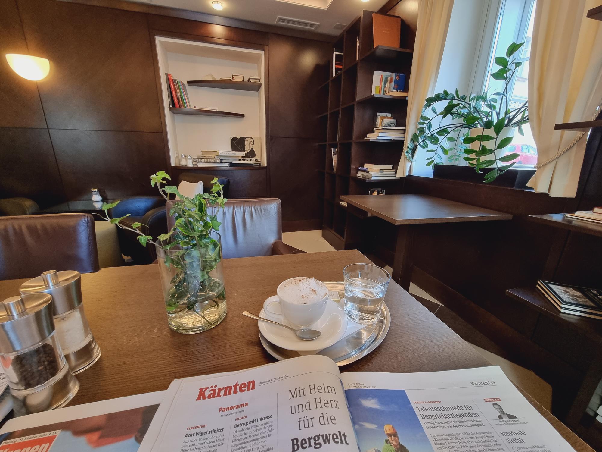 Die Bücherlounge im Cafe Sandwirth mit Bücherregalen und großer Zeitschriftenauswahl, hier mit einem Capuccino am Tisch sowie einer aufgeblätterten Tageszeitung