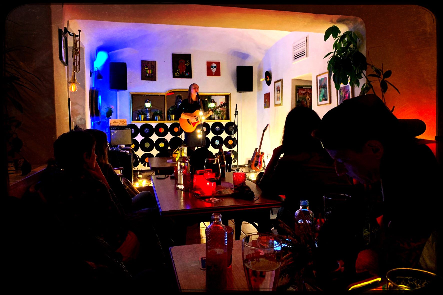 Eine junge Musikerin mit Akustikgitarre steht auf der kleinen Bühne im Wohnzimmer und singt ins Mikrofon, während das Publikum ihr lauscht