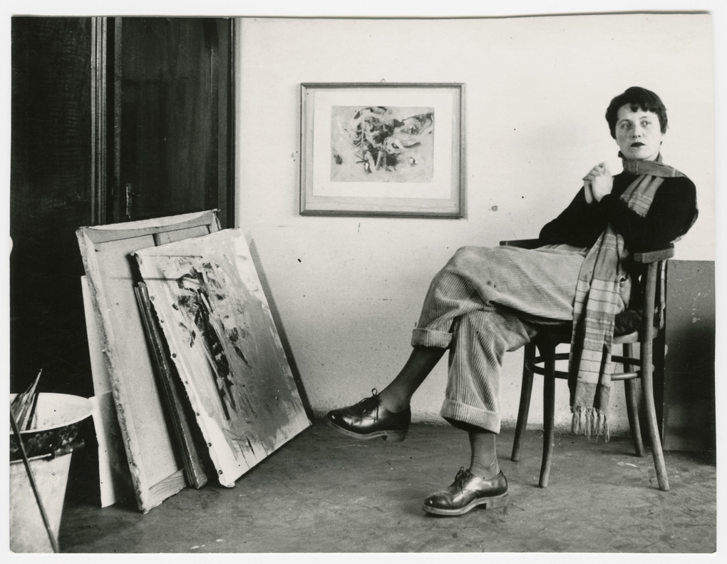 Bild von Maria Lassnig, die vor ihren Werken im Atelier sitzt