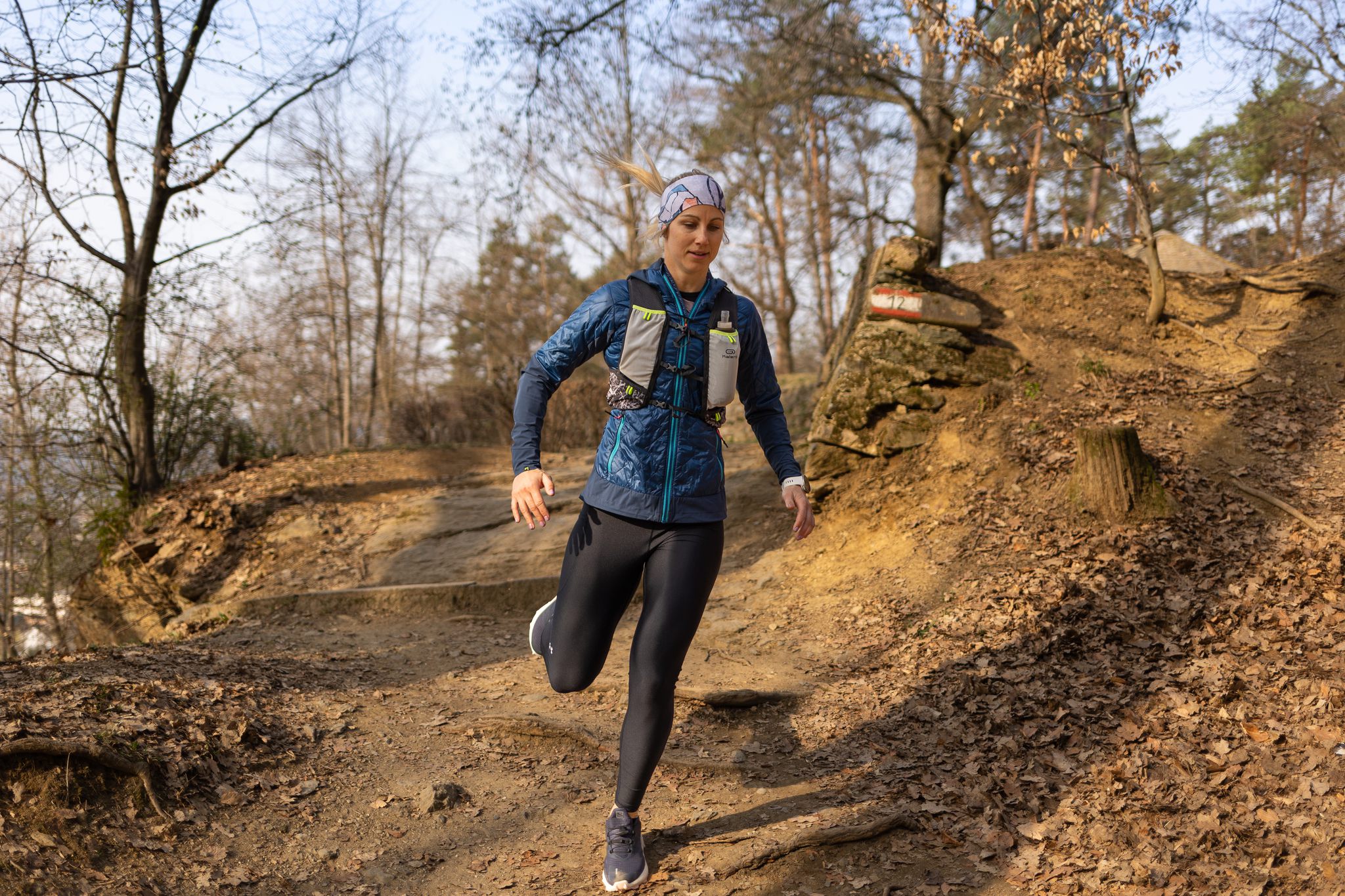 Trail Läuferin in schwarzer Sportlegging dunkelblauer Laufjacke und Stirnband beim Laufen auf Waldweg 