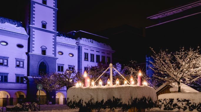 Ein überdimensionaler, mit vielen Kerzen erleuchteter Adventskranz vorm verschneiten Domplatz in Klagnefurt