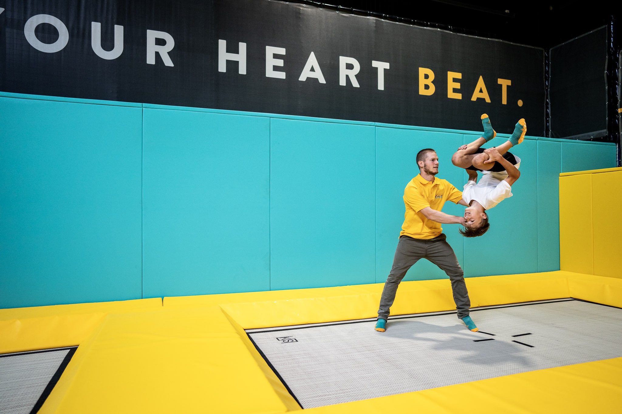 Ein zertifizierter Trampolintrainer hilft einem Kind auf einem Bodentrampolin beim Üben eines Vorwärtssaltos