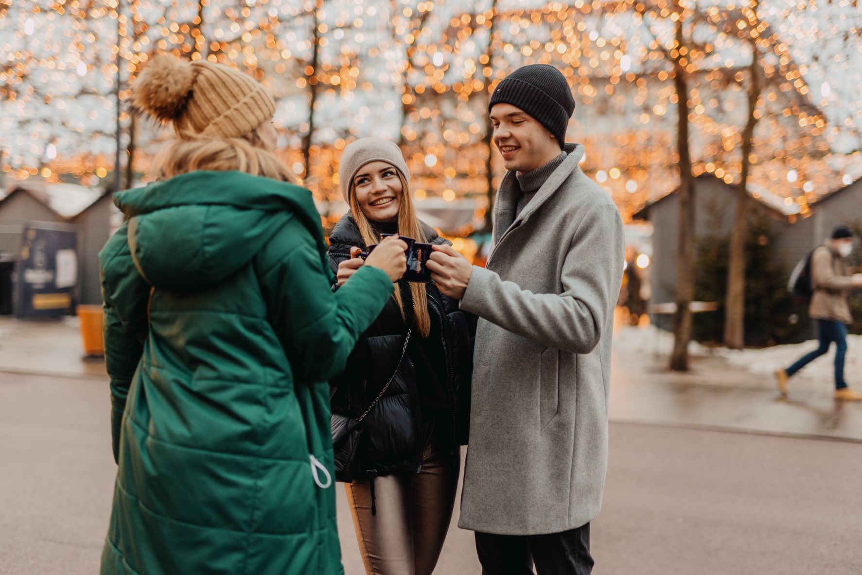 Zwei junge Frauen und ein junger Mann stoßen am Christkindlmarkt Klagenfurt mit Glühwein an. Im Hintergrund Weihnachtsbeleuchtung und Markthütten