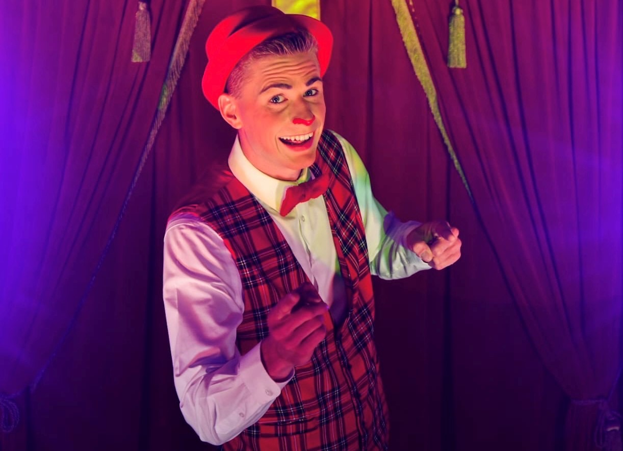 Der junge Zirkusdirektor vom Circus Dimitri mit rot-kariertem Gilet, roter Masche, rotem Hut und roter Nase lächelt in die Kamera