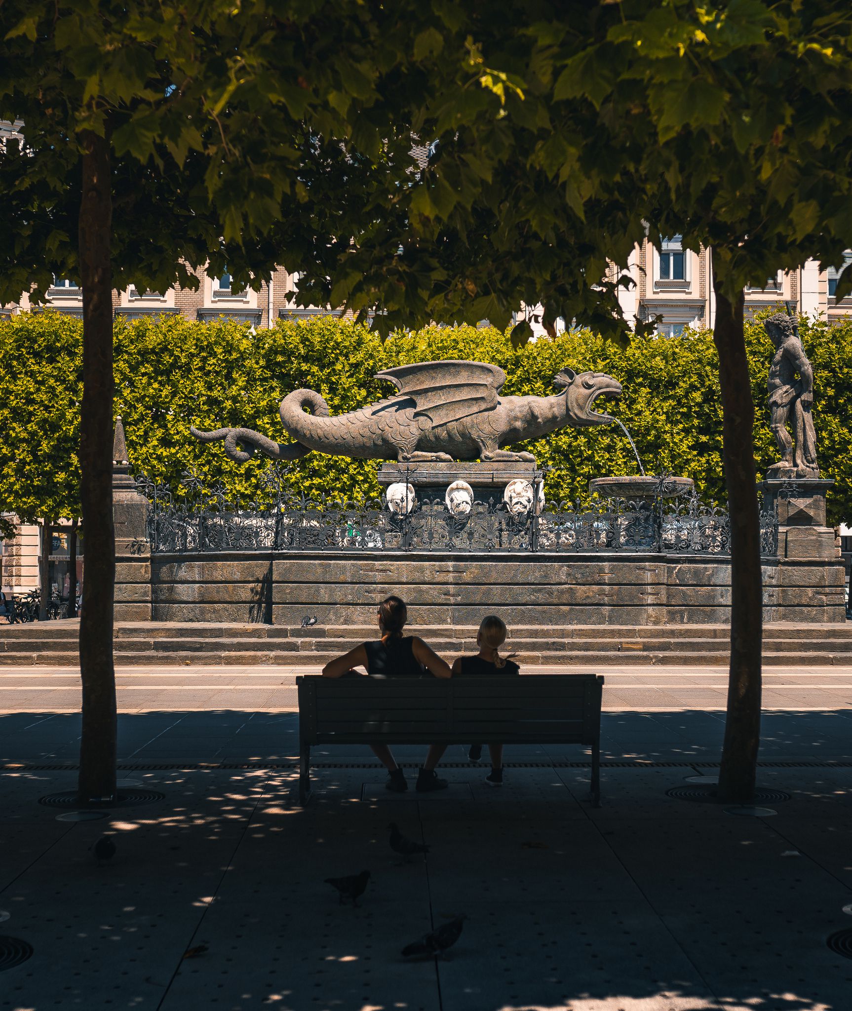Bild von zwei Personen, die auf einer Parkbank sitzen mit Blick auf den Lindwurm von der Seite