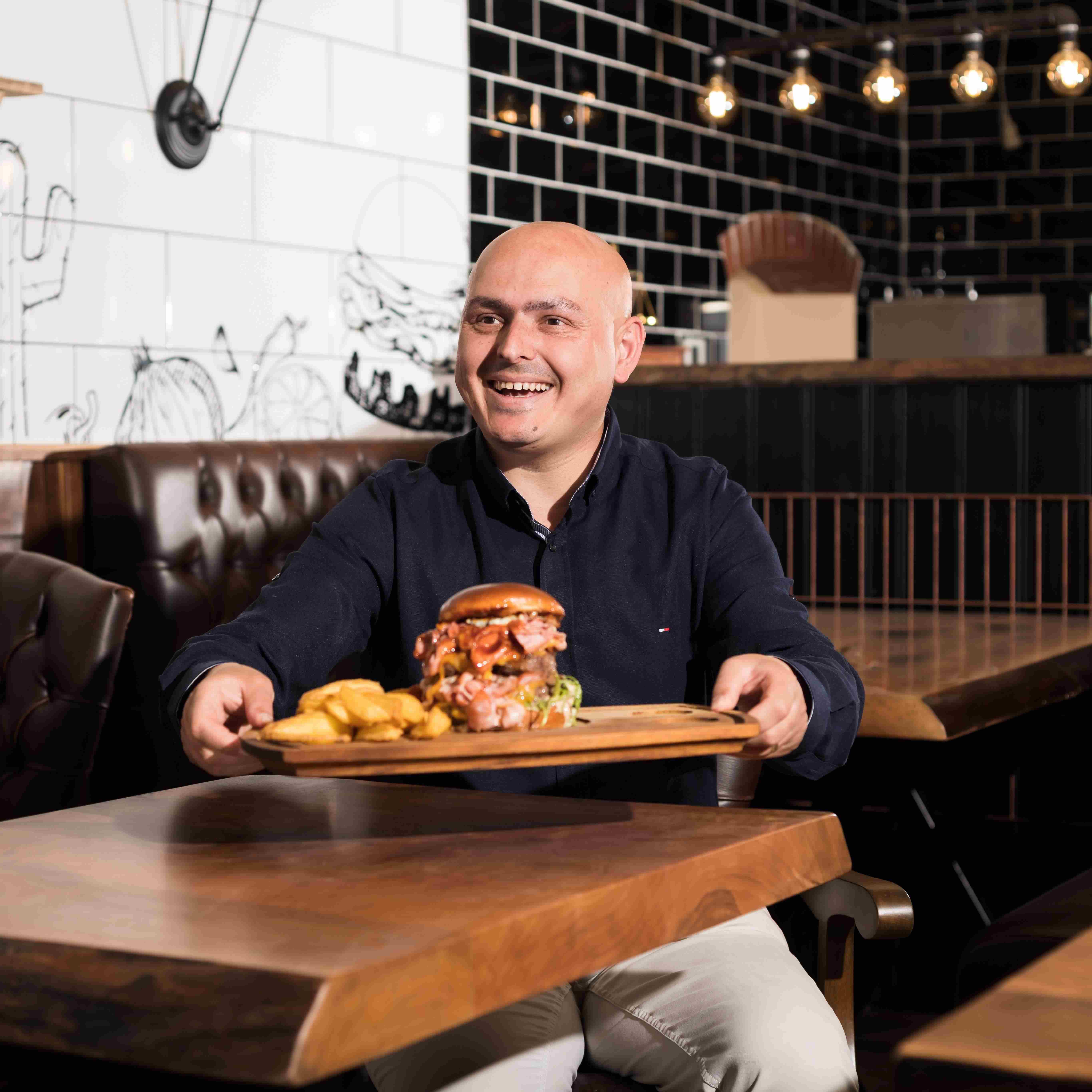 Burger Boutique Chef Ümit Baran an einem seiner Tische mit einem Tablet, auf dem ein Burger steht, in der Hand