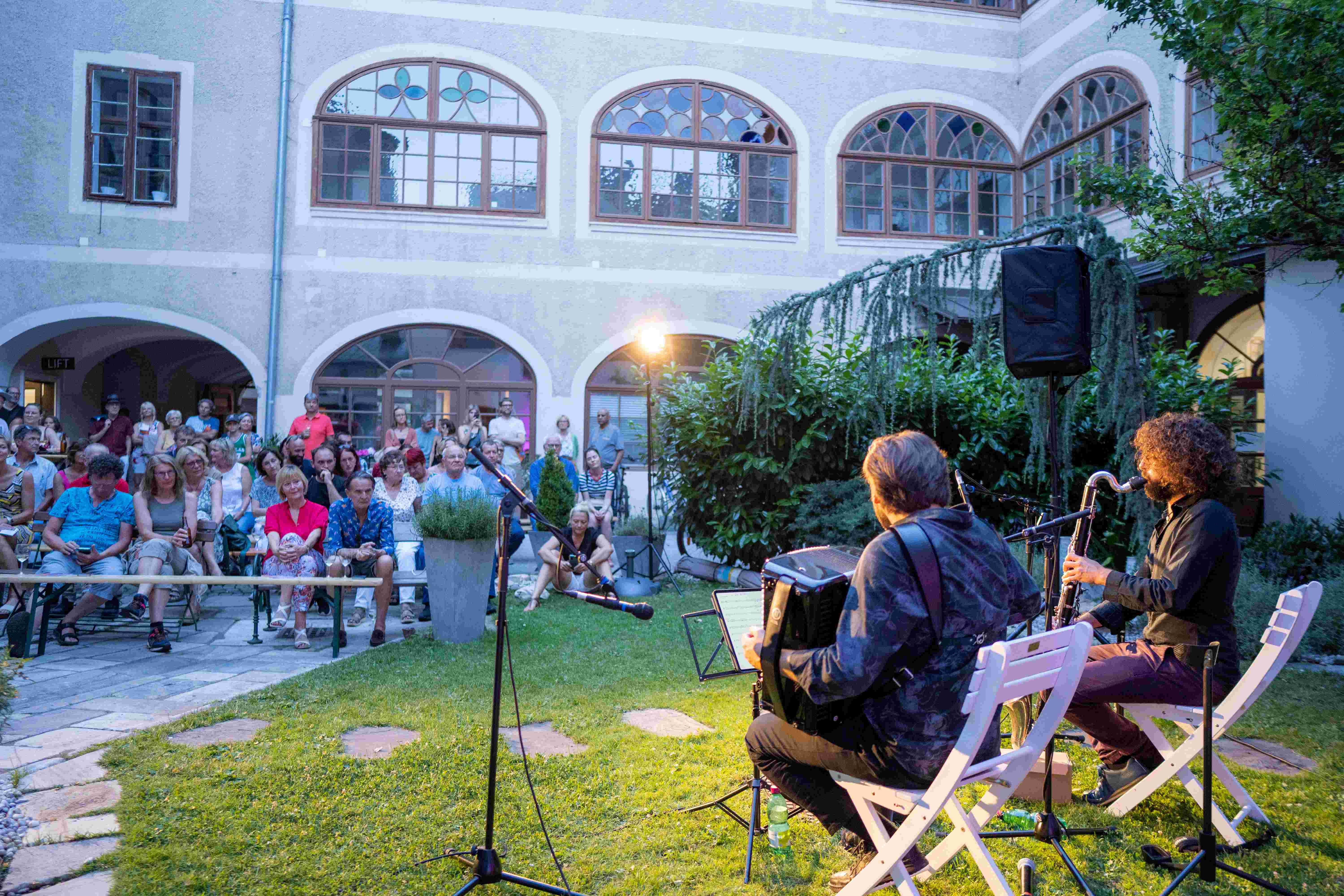 Donnerszenen Foto aus dem Innenhof des Ossiacherhofes mit zwei Musikern im Vordergrund, die vor sitzendem Publikum in der beginnenden Dämmerung spielen
