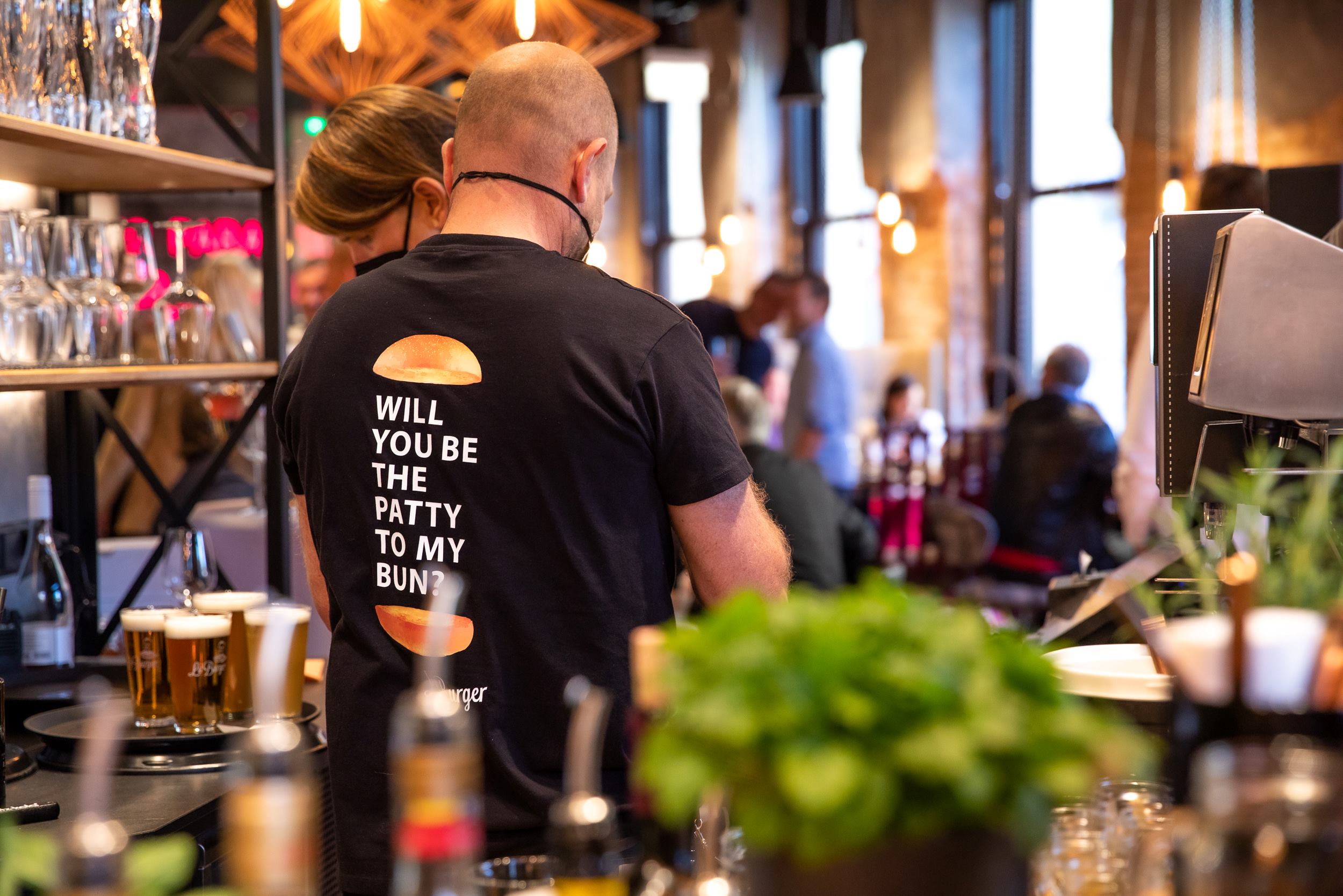 Szene an der Bar von Le Burger Klagenfurt, ein Mann trägt die Aufschrift "Will you be the patty on my bun" am T-Shirt-Rücken