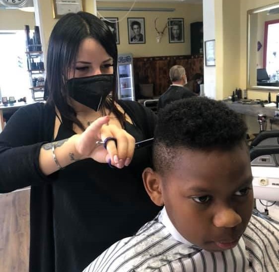 Eine Barbiere des Barbershop Glaser steht hinter einem jungen Mann und schneidet seine Haare am Hinterkopf