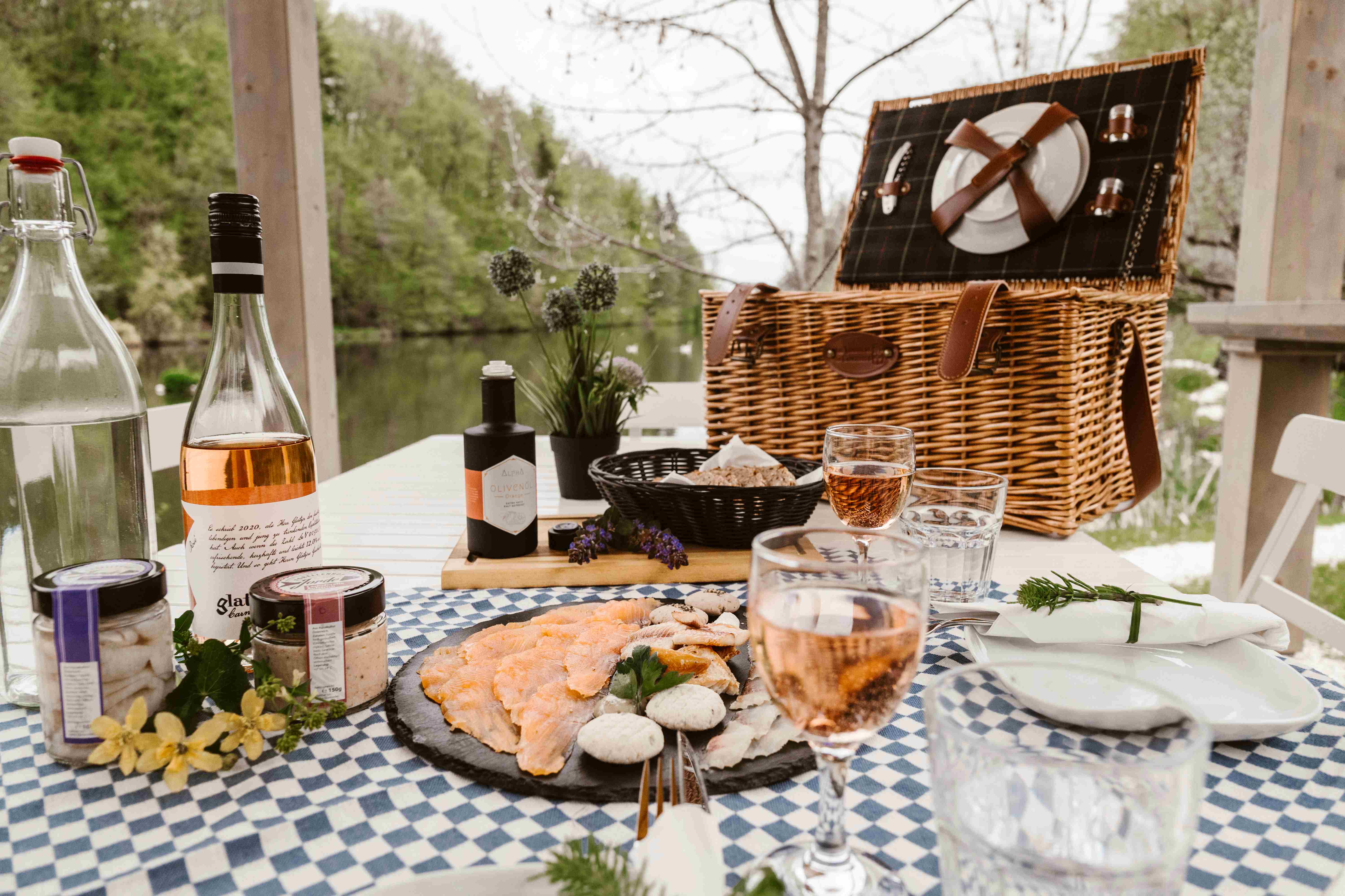Ein Teller voller Fisch-Spezialitäten auf karierter Tischdecke mit Picknickkorb und Fischteich im Hintergrund