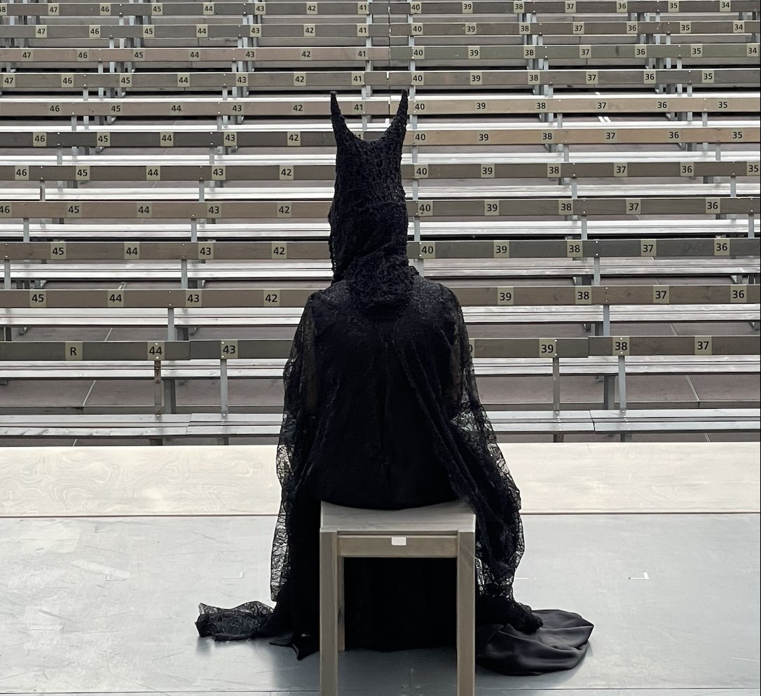 Zu sehen ist die Jedermann-Figur des Teufels von hinten, sitzend auf einem Stuhl auf der leeren Bühne vor leeren Besucherrängen