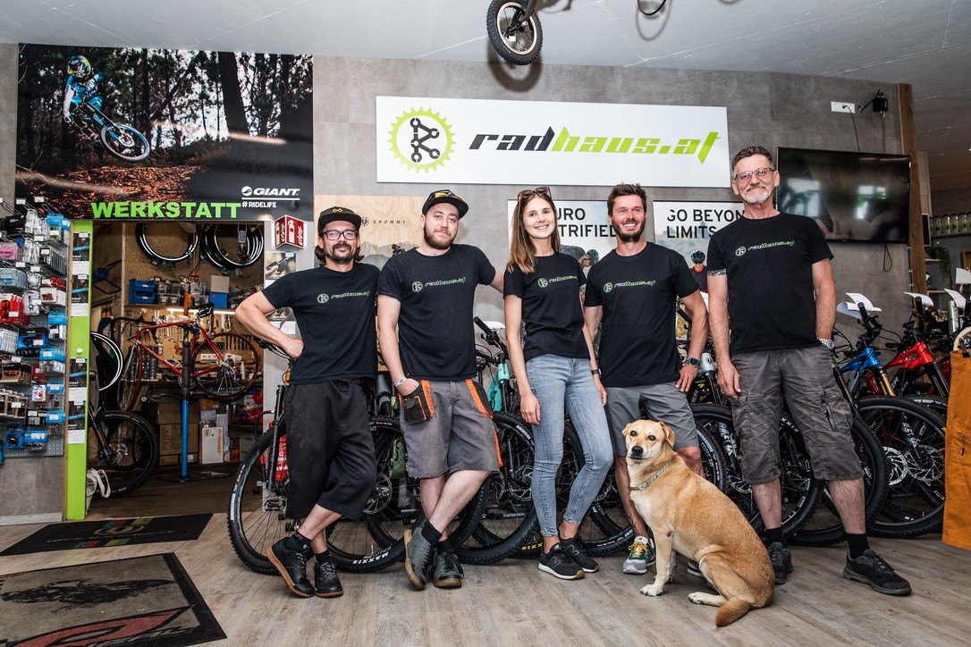 Fünf Teammitglieder des Klagenfurter Fahrrad-Shops Radhaus stehen im Verkaufsraum mit dem Logo im Hintergrund und einem braunen Hund im Vordergrund