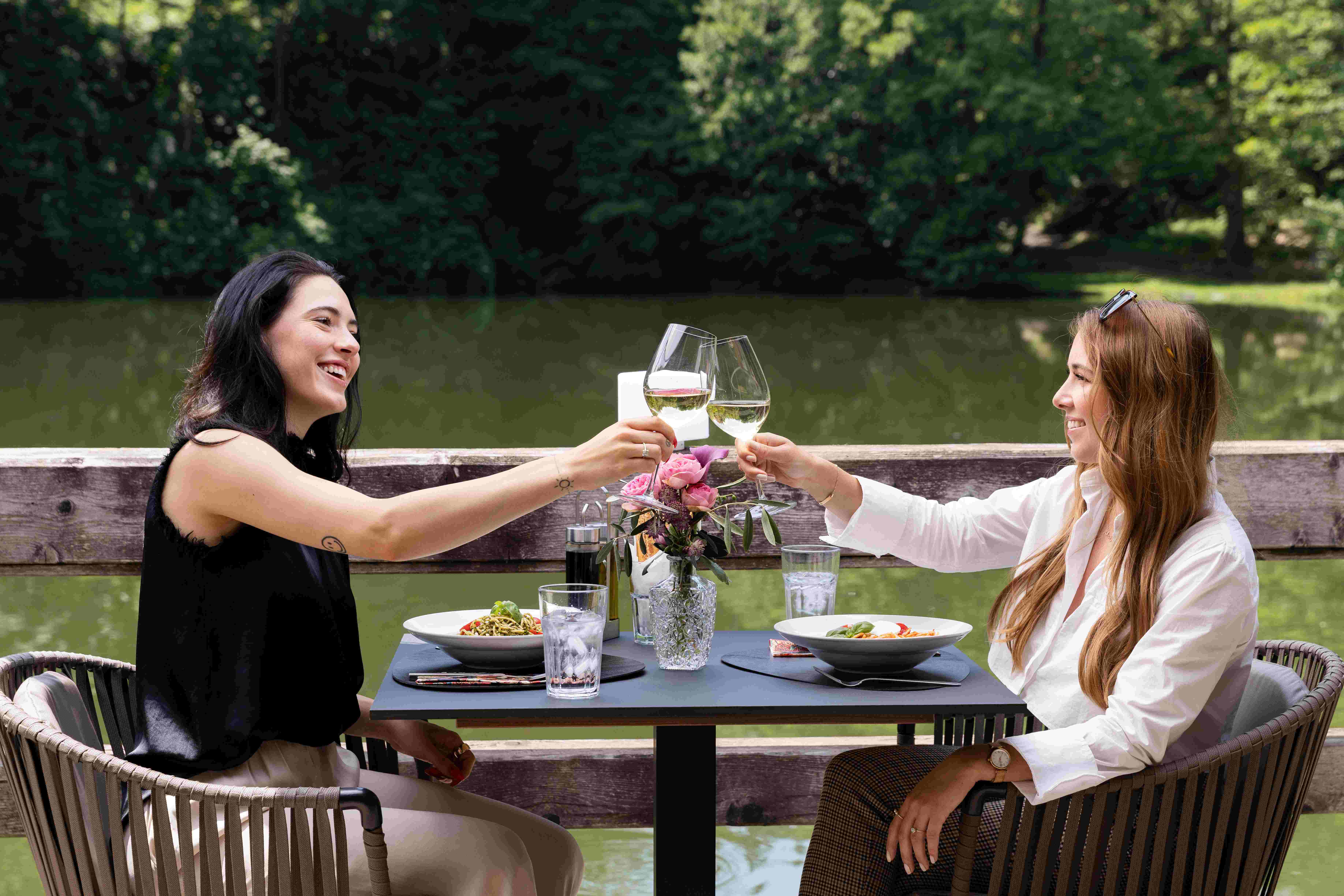 Zwei Frauen stoßen mit Wein an. Sie sitzen auf der REstaurantterrasse am Teich des Fischerwirt Conamore in Klagenfurt